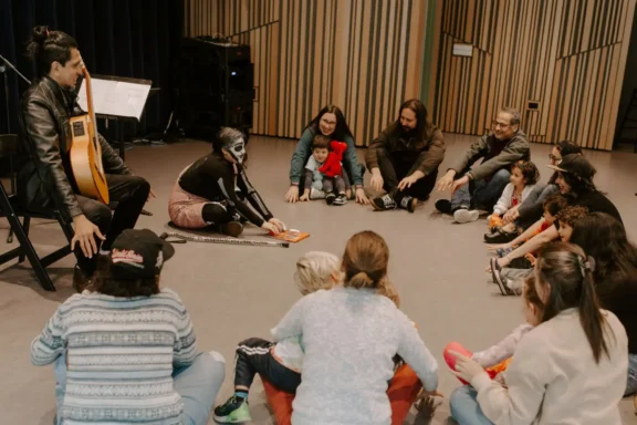 A group of adults and children sit in a circle on the floor, listening to a person in face paint read a book, with a guitarist seated nearby.