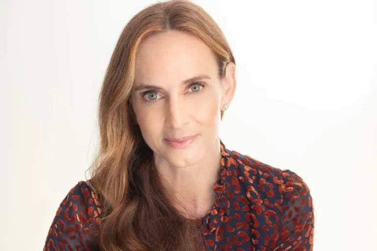 A woman with long, wavy brown hair and light eyes looks at the camera, wearing a patterned red and black top against a plain light background—her confident gaze embodying the spirit of The 26/27 Goodman Education Benefit.