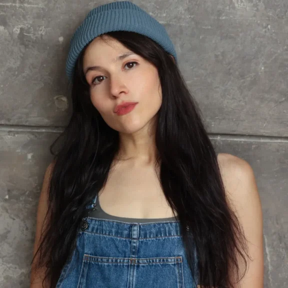 Woman with long dark hair wears a blue knit beanie and denim overalls, standing in front of a gray textured wall, looking directly at the camera with a neutral expression.