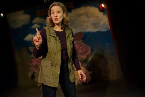 A woman with shoulder-length hair gestures with her index finger raised while speaking on stage, in front of a painted backdrop with clouds and flowers.