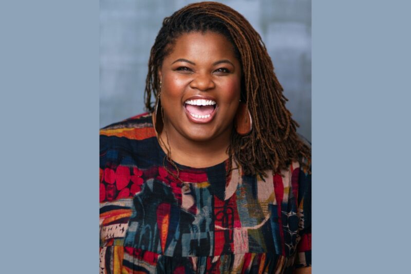 A woman with long, brown, braided hair and wooden earrings is smiling widely. She is wearing a colorful patterned shirt and is pictured in front of a plain, gray backdrop.