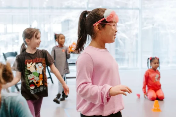 Children participate in an indoor group activity, smiling and moving around a brightly lit room with large windows in the background.