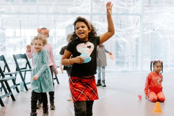 Children play an indoor game; one girl in a plaid skirt and heart shirt raises her hand while others watch or participate. Bright windows and chairs are in the background.