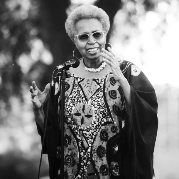 An older woman in patterned clothing and glasses speaks and gestures at a microphone outdoors.