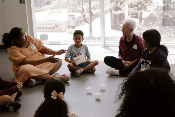A group of children and adults sit in a circle on the floor indoors, talking. Three empty plastic cups are placed in the center of the circle.