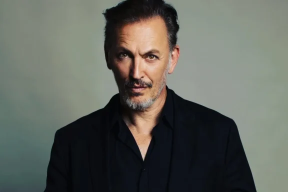 A middle-aged man with short dark hair and a trimmed beard wears a black collared shirt and black jacket, looking directly at the camera against a neutral background.