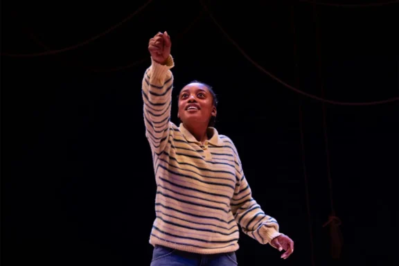 A person in a striped sweater stands on stage with one arm raised, looking upward, against a dark background with visible ropes.