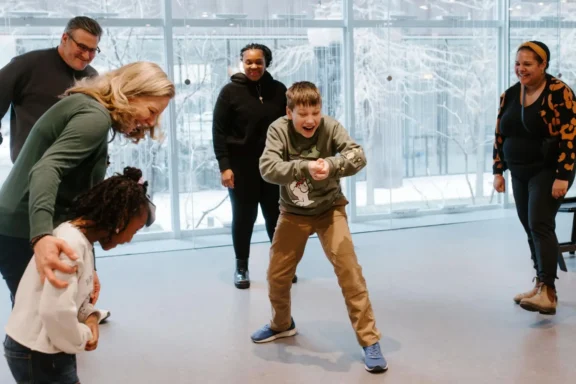 Five people stand indoors on a gray floor while one boy in the center enthusiastically mimics a dance move. Snowy trees are visible through large windows in the background.