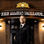 A man in a suit stands in front of a building with a sign reading “Dennis Watkins The Magic Parlour” above the entrance.