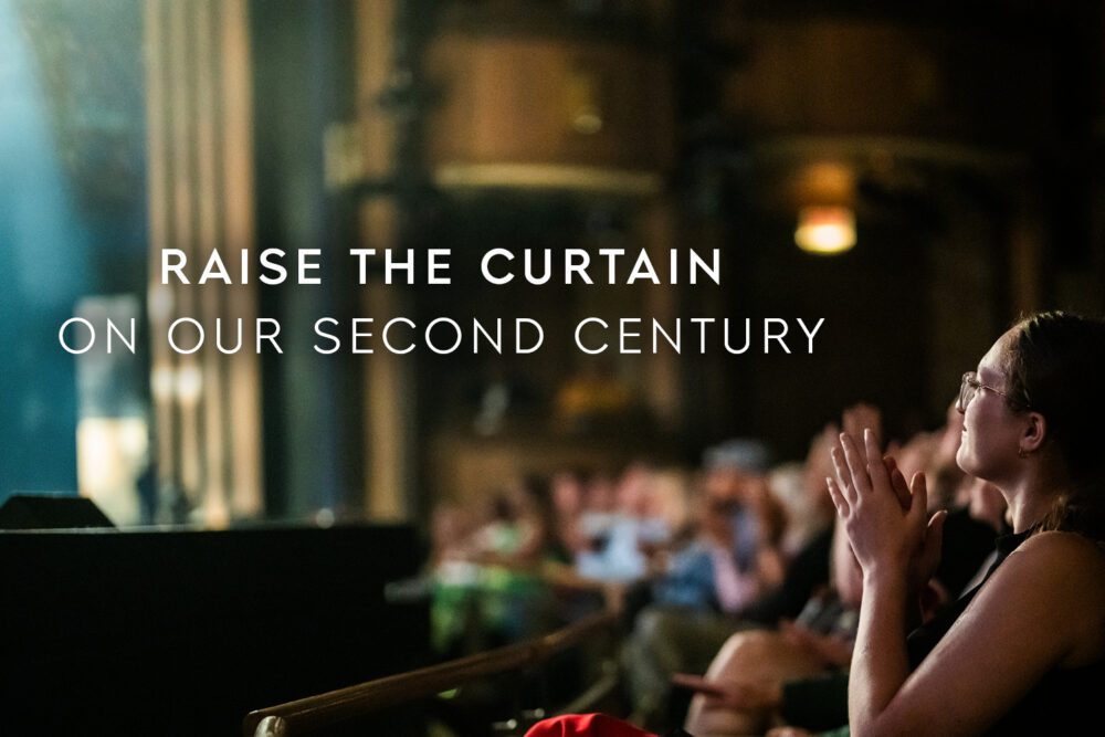 A woman applauds in a theater audience as a spotlight shines on stage, with text reading “Raise the curtain on our second century.”.