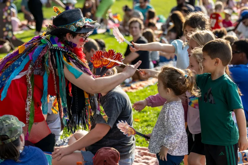 A performer in colorful attire interacts with a group of young children holding flower props at an outdoor event with a seated audience.