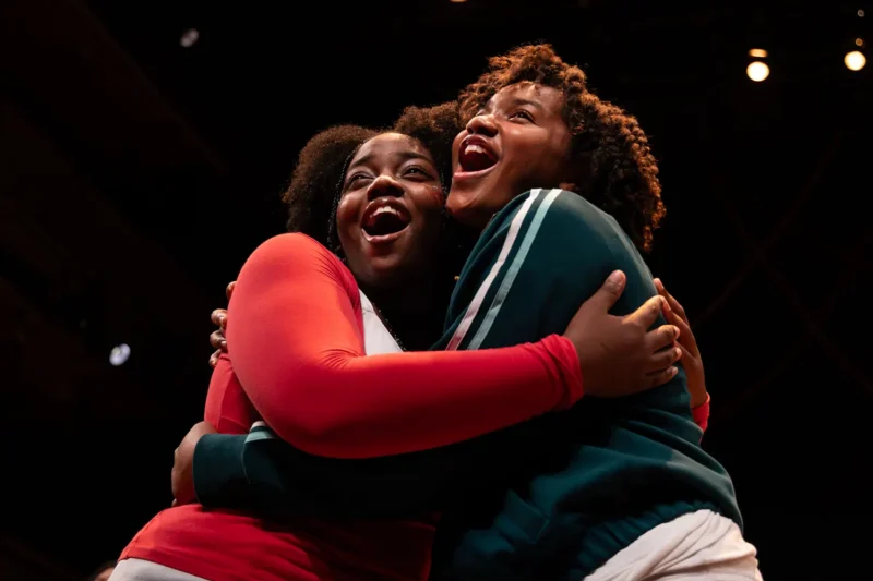 Two people stand on stage, smiling and hugging each other under stage lights, appearing joyful and enthusiastic.