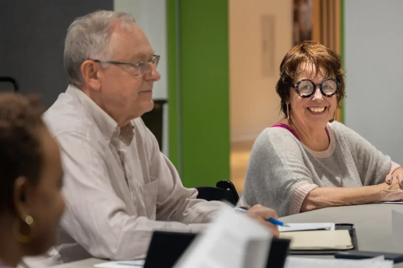 Two older adults sit at a table with papers and notebooks. The man looks forward, while the woman smiles and wears glasses. Another person is partially visible in the foreground.
