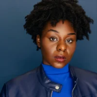 A person with short curly hair wears a blue turtleneck and blue jacket, posing against a solid blue background with a neutral expression.