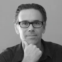 A man with dark-rimmed glasses and short hair sits facing the camera, resting his chin on his hand, wearing a dark collared shirt, against a plain background.