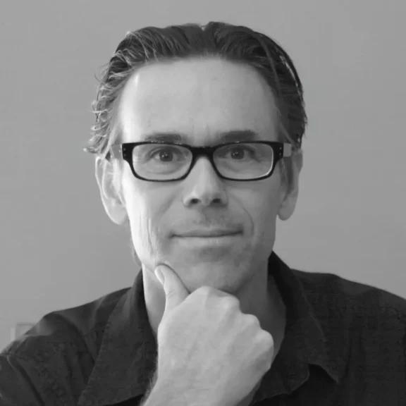 A man with dark-rimmed glasses and short hair sits facing the camera, resting his chin on his hand, wearing a dark collared shirt, against a plain background.