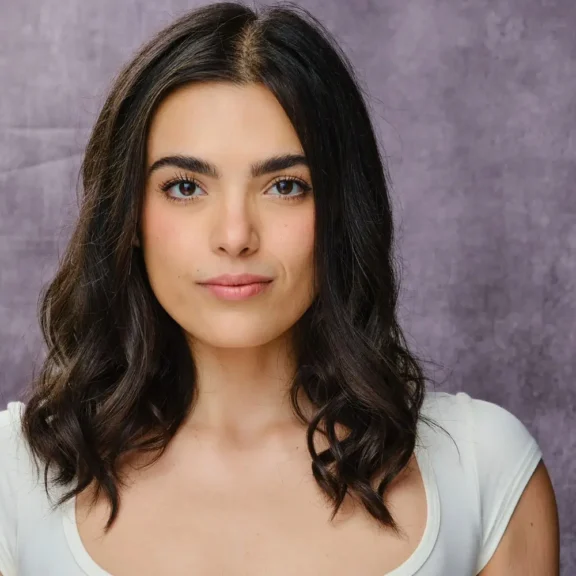Woman with wavy dark hair wearing a white top, standing in front of a textured gray background, looking directly at the camera with a neutral expression.