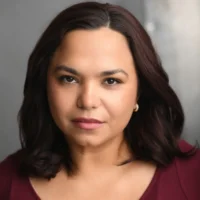 A person with shoulder-length dark hair and gold hoop earrings looks directly at the camera with a neutral expression, against a gray background.