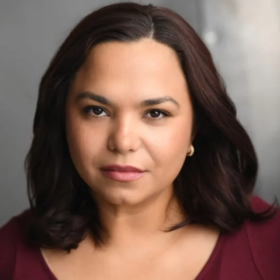 A person with shoulder-length dark hair and gold hoop earrings looks directly at the camera with a neutral expression, against a gray background.