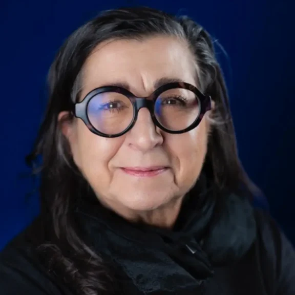 A woman with long dark hair, wearing large round black glasses and a black scarf, poses in front of a dark blue background.