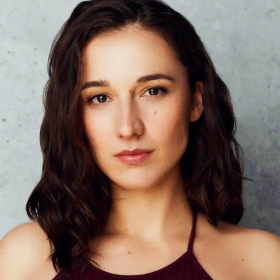 A woman with shoulder-length brown hair and a maroon top looks directly at the camera against a gray background.