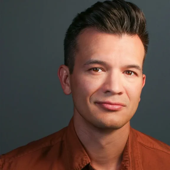 A man with short, dark hair and a slight smile wears a rust-colored collared shirt against a plain dark background.