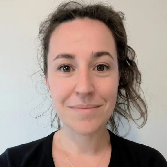A woman with wavy brown hair pulled back, wearing a black top and a necklace, stands in front of a plain light-colored wall, looking directly at the camera with a neutral expression.