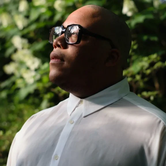 A person wearing large black glasses and a white button-up shirt looks upward, standing outdoors with greenery and blurred white flowers in the background.