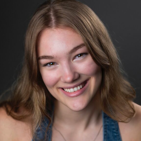 Young woman with long light brown hair smiling at the camera, wearing a blue top, with a neutral dark background.