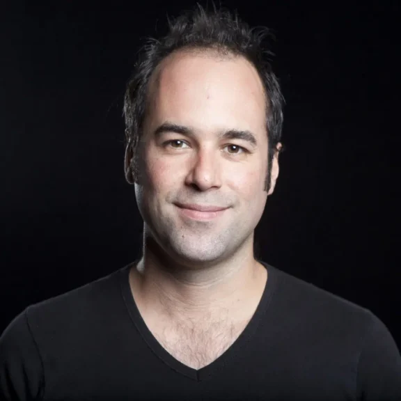 A man with short dark hair and a receding hairline wearing a black V-neck shirt stands against a plain black background, smiling slightly at the camera.