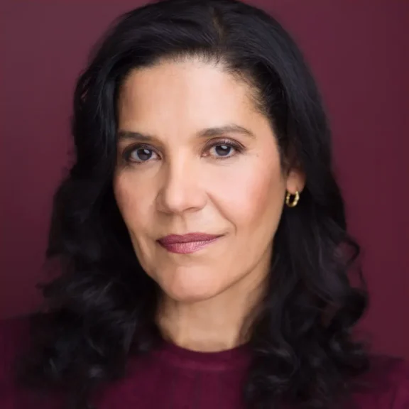 Woman with medium skin tone and wavy dark hair, wearing gold hoop earrings and a burgundy top, looks calmly at the camera against a matching burgundy background.