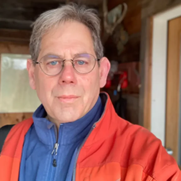 Middle-aged man with glasses and gray hair wearing an orange jacket over a blue zip-up, standing indoors with wooden walls and a door in the background.