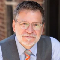 A middle-aged man with short gray hair, beard, and glasses, wearing a gray vest, light blue shirt, and orange-striped tie, sits in front of a brick wall background.