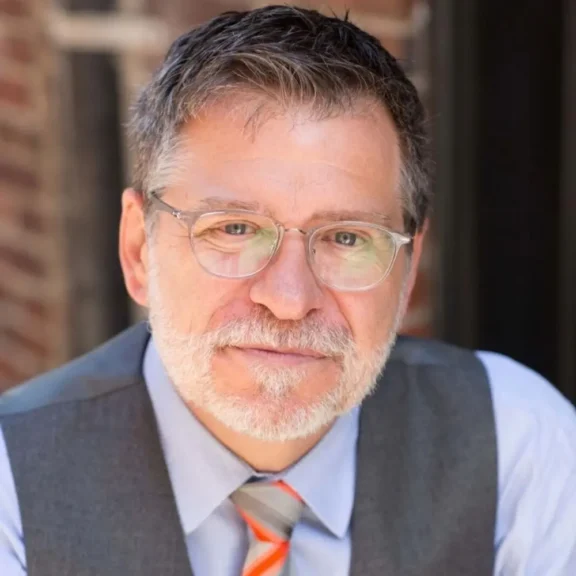 A middle-aged man with short gray hair, beard, and glasses, wearing a gray vest, light blue shirt, and orange-striped tie, sits in front of a brick wall background.
