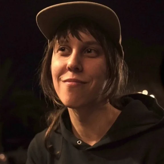 A person wearing a baseball cap and hoodie sits in a dimly lit setting, looking slightly upward with a faint smile.