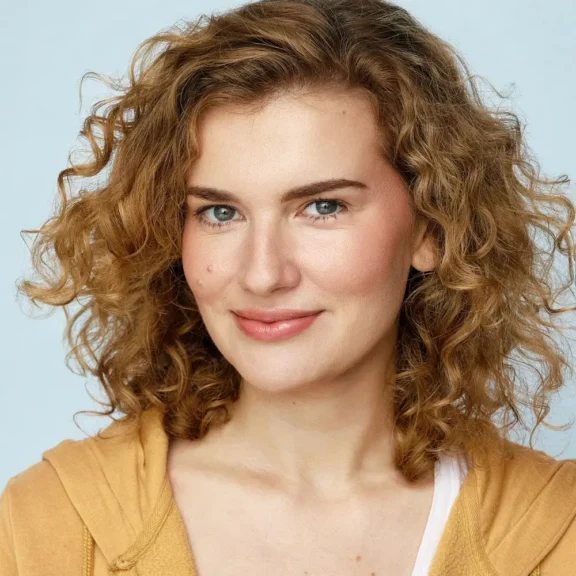 A person with curly light brown hair, blue eyes, and light skin wears a yellow hoodie and smiles at the camera against a light blue background.