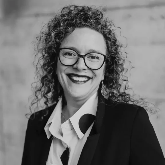 A woman with curly hair and glasses smiles, wearing a suit jacket, patterned shirt, and earrings, standing in front of a plain background.