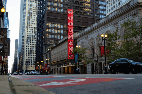 Street view of the Goodman Theatre with a red vertical sign, surrounded by tall buildings, cars, and streetlights in an urban setting.