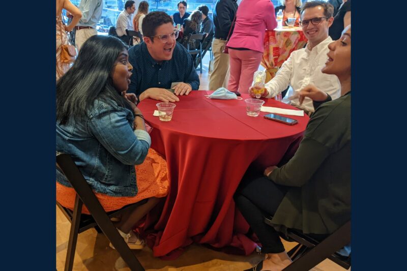 Four people sit around a red tablecloth-covered table, talking and laughing at an indoor social gathering with drinks and food nearby.