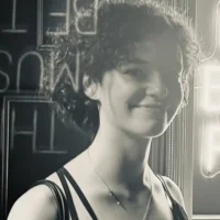 A woman with curly hair smiles at the camera in front of a neon sign and a dark wall. The photo is in black and white.