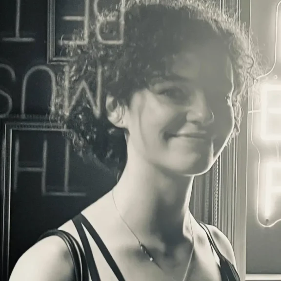 A woman with curly hair smiles at the camera in front of a neon sign and a dark wall. The photo is in black and white.