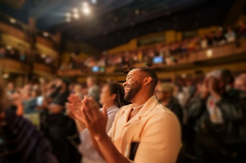 A man stands and applauds in a crowded theater, surrounded by people who are also standing and clapping. The atmosphere appears lively and celebratory.