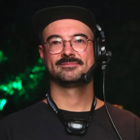 A person with glasses, a headset, and a black cap stands against a dark background with green light.
