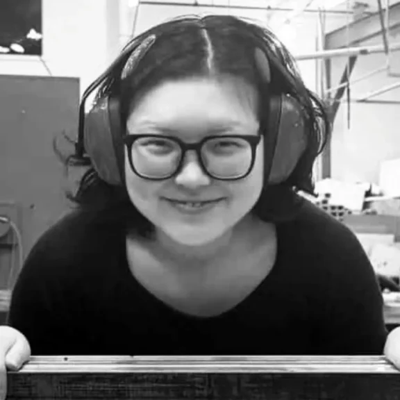 Person with glasses and earmuffs smiles while working in an industrial setting, holding a wooden object. Black and white image.