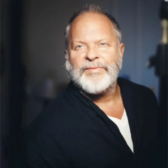 A middle-aged man with a gray beard and short hair wears a dark jacket over a white shirt, standing indoors with a soft, neutral background.