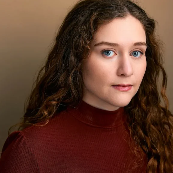 A woman with long curly brown hair and blue eyes wearing a maroon turtleneck top looks at the camera against a neutral background.