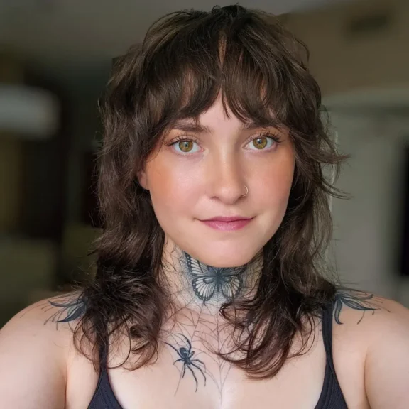 Person with wavy brown hair, floral and spider tattoos on neck and chest, wearing a nose ring and a black top, looking at the camera.