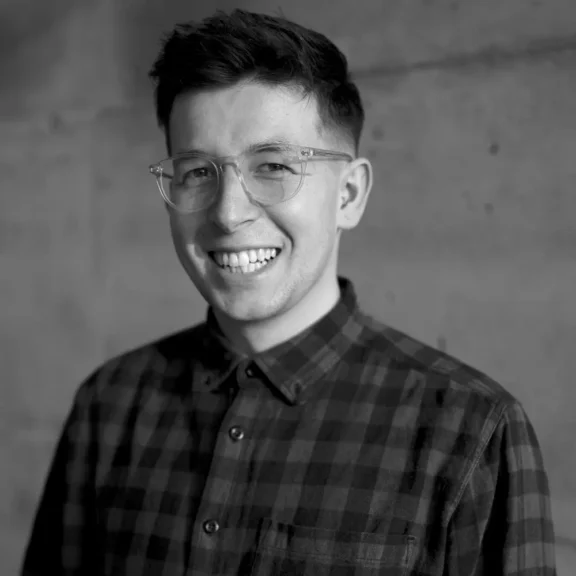 A person with short hair and glasses, wearing a plaid shirt, smiles while standing in front of a plain concrete wall.