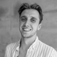 A young man with short, wavy hair and a light beard, wearing a striped shirt, smiles at the camera against a plain, neutral background.