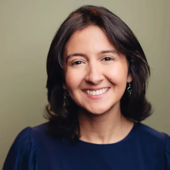A person with medium-length dark hair, wearing green earrings and a navy blue top, smiles at the camera against a plain light background.